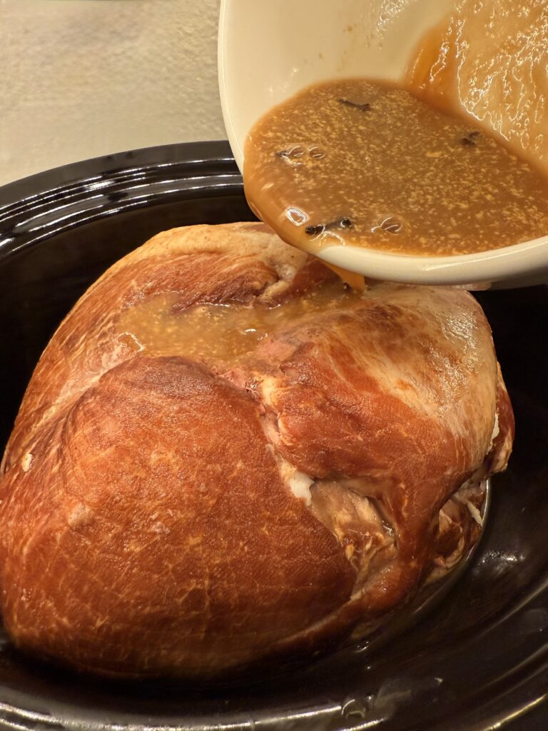 Pouring juice, sugar, honey, cloves and mustard mixture over ham in slow cooker on the counter.