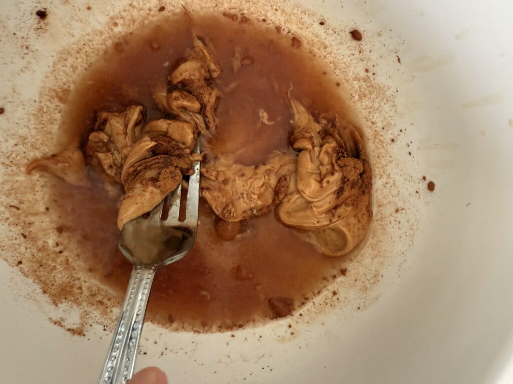 apple juice, vanilla, cardamon, cinnamon and peanut butter in a bowl being whisked by a fork.