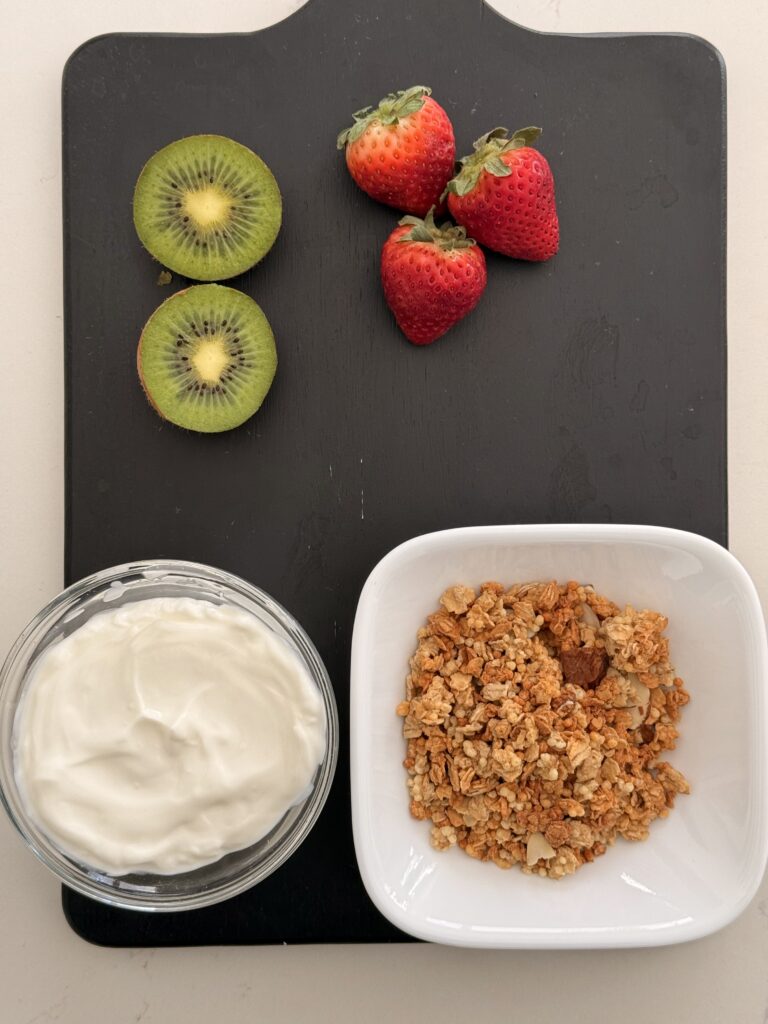 Greek yogurt in a clear bowl, granola in a small white bowl and a cut in half kiwi fruit next to 3 whole strawberries on a black cutting board on the counter.