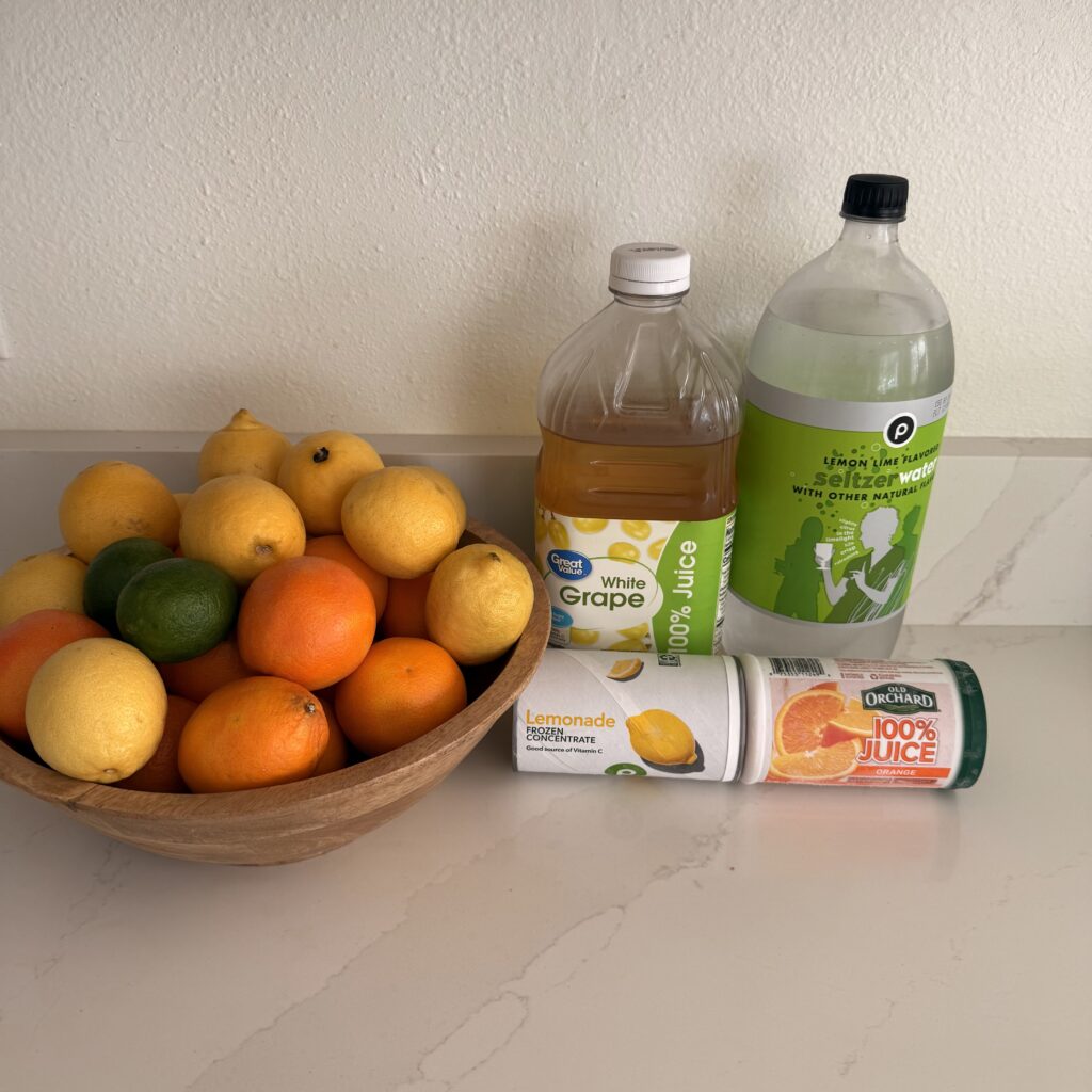 Bowl of oranges, limes and lemons in a bowl on the counter with white grape juice, lime seltzer water, frozen lemonade concentrate and frozen orange juice concentrate on the counter.