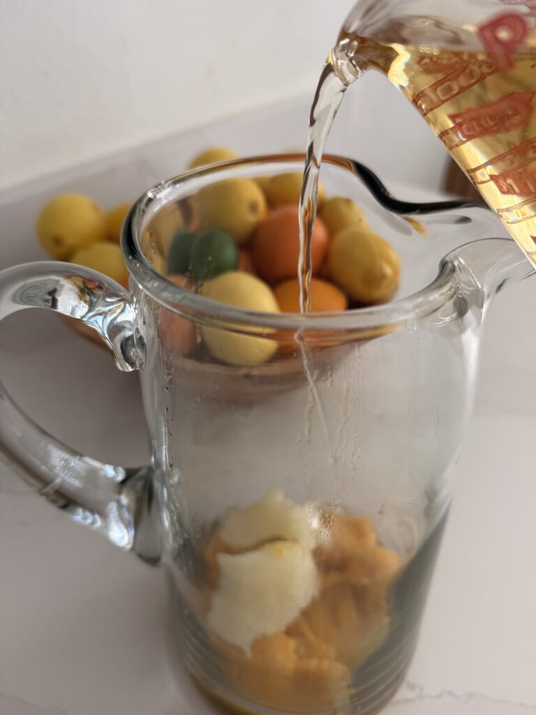 Pouring white grape juice into a glass pitcher with frozen fruit concentrate on the counter. a bowl of citrus fruit in the background on the counter.