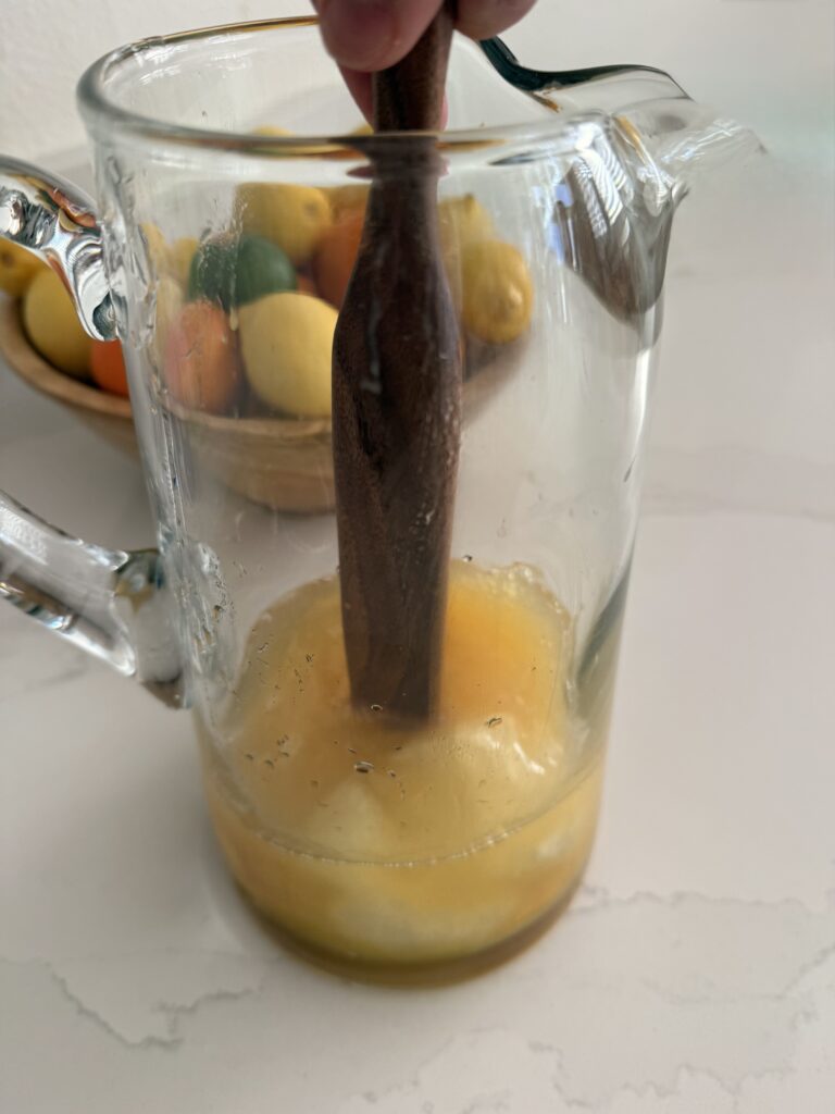 mixing the frozen juice concentrates with the white grape juice in a glass pitcher on the counter. a bowl of citrus fruit in the background on the counter.