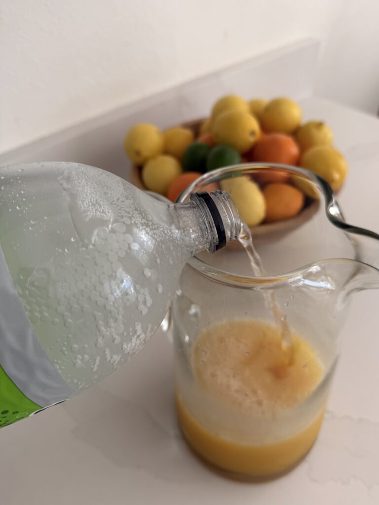 Pouring lime seltzer water into a glass pitcher with juices in it on the counter. A bowl of citrus fruit in the background on the counter.