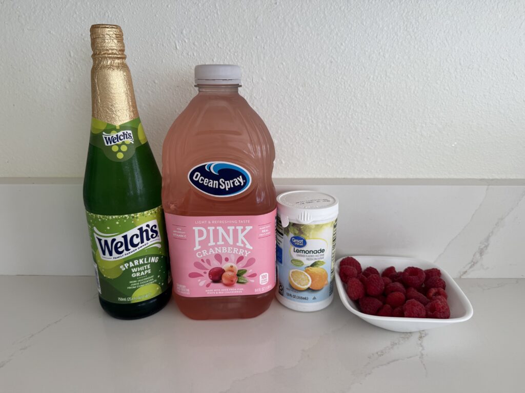 sparkling white grape juice, pink cranberry juice, frozen lemonade concentrate and fresh raspberries in a white bowl on the counter.