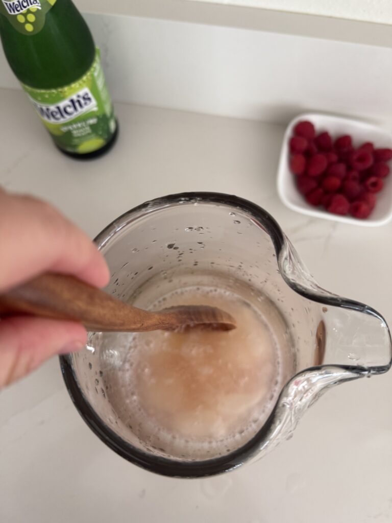 Frozen lemonade concentrate and pink cranberry juice in a glass pitcher being mixed with a wooden spoon on the counter. In the background is fresh raspberries in a small white bowl and a bottle of sparkling white grape juice on the counter.