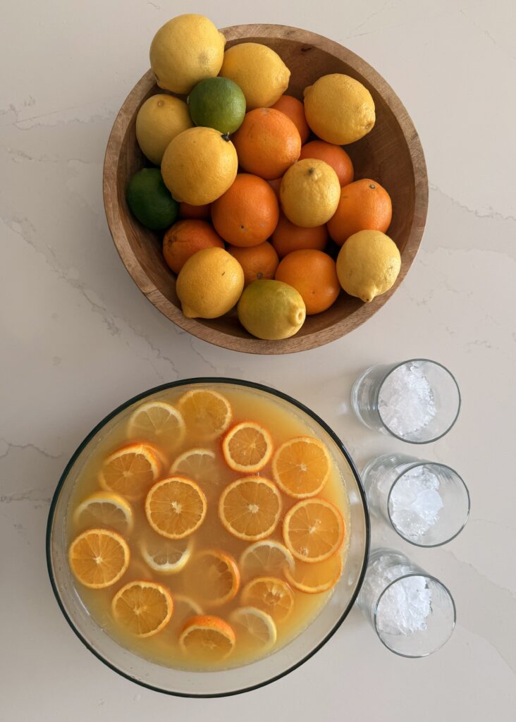 Citrus Fruit Party Punch in a punch bowl and on the counter. A bowl of citrus fruit in a brown bowl on the counter beside the punch and three ice filled punch glasses on the counter.