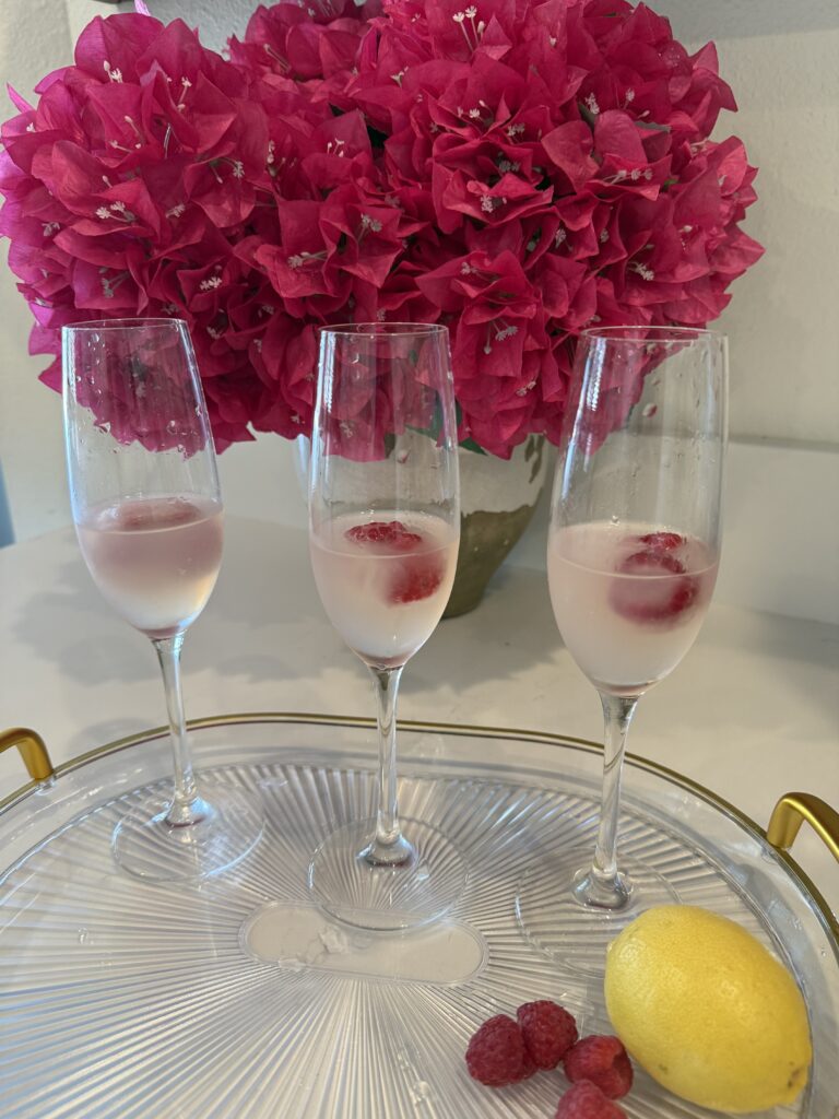 three champagne glasses with reserved juice and raspberry ice cubes in them on a glass tray on the counter. Pink flowers in a vase in the background. Lemon and raspberries on the tray on the counters.