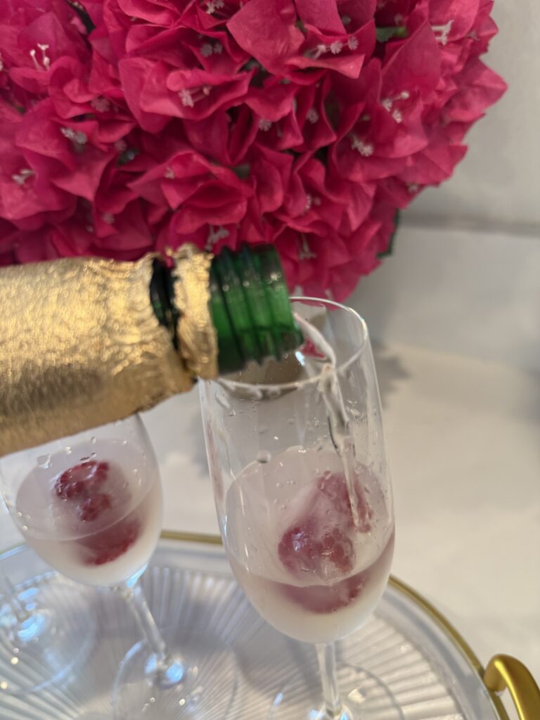 pouring sparkling white grape juice into a champagne flute on a glass tray on the counter.  Pink flowers in a vase in the background on the counter.