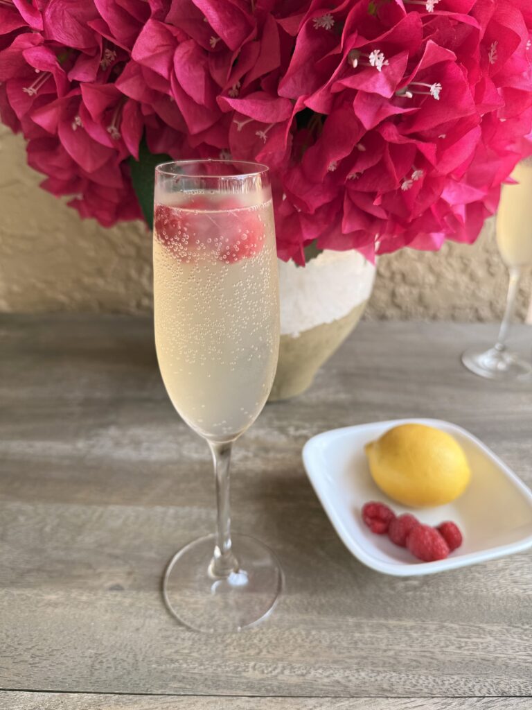 Bubbly Berry Mocktail with pink flowers on a vase and a lemon and raspberries in a small white dish on a brown outside table.