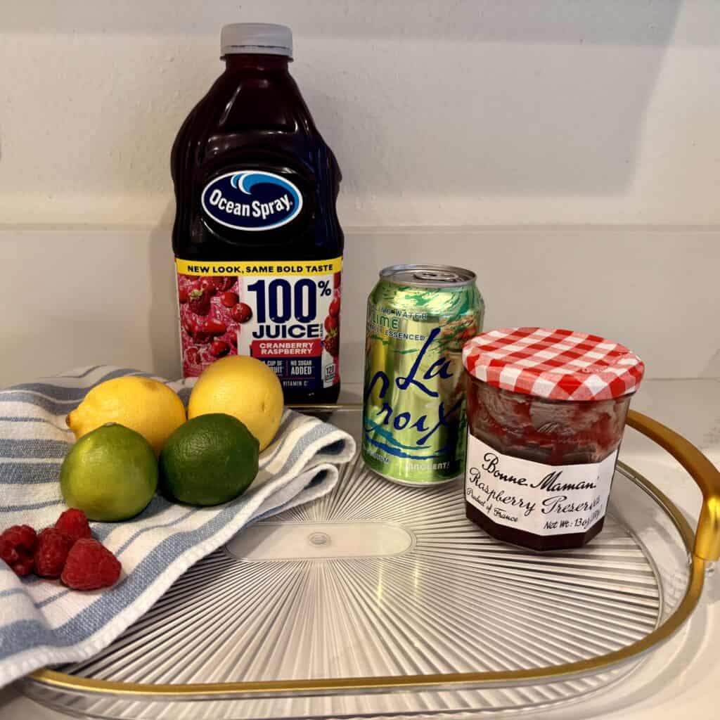 Cranberry raspberry juice, lime seltzer water, raspberry jam, lemons and limes and raspberries on a glass tray with a blue and white dish towel on the counter.