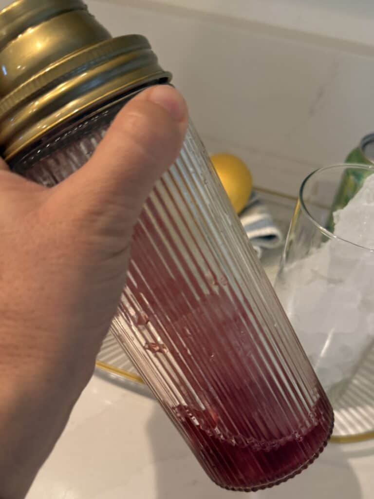 Cocktail shaker with raspberry jam, ice and cranberry raspberry juice in it being held in the air my left hand. Glass tray with tall glass on glass tray on the counter in the background.