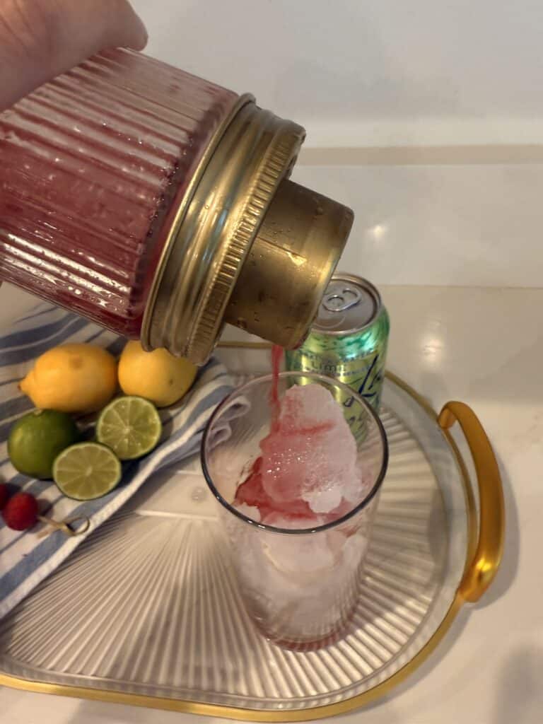 Cocktail shaker straining ingredients into a tall ice filled glass on a glass tray on the counter.