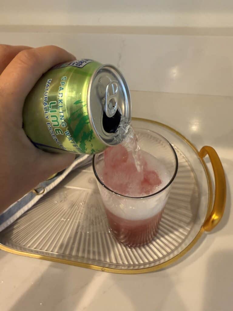 Lime seltzer water being poured in a tall glass on a glass tray on the counter.