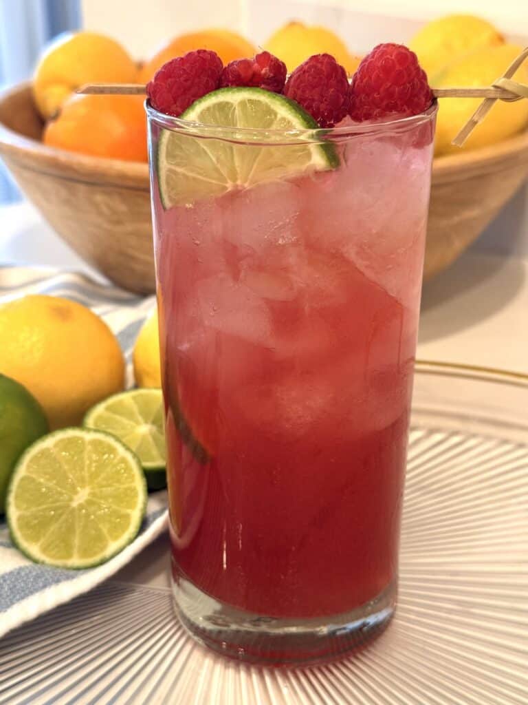 Cran-Raspberry Lime Rickey on a glass tray on the counter surrounded by by citrus fruit.