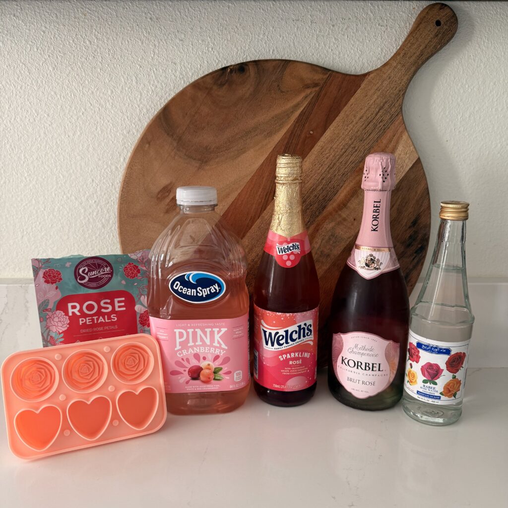 rose ice cube mold, edible rose petals, Pink Cranberry Juice,Rose water (food grade), Rose Prosecco and Sparkling Rose non-alcoholic Grape Juice Cocktail on the counter.