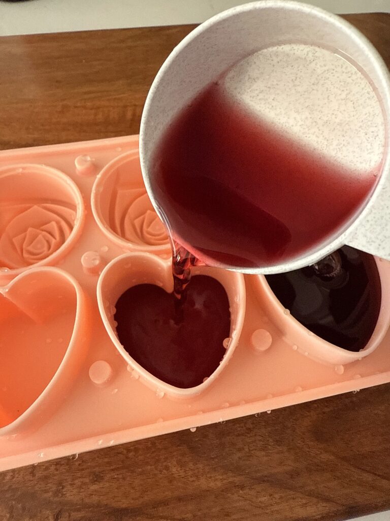 pouring cran-raspberry juice into hear shaped ice cube molds on a brown cutting board.