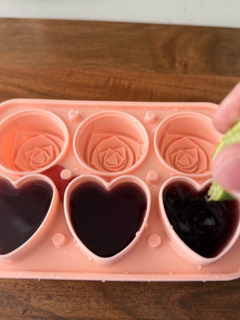 squeezing a lime and its juice into heat shaped ice cube mold filled with cran-raspberry juice on a brown cutting board.