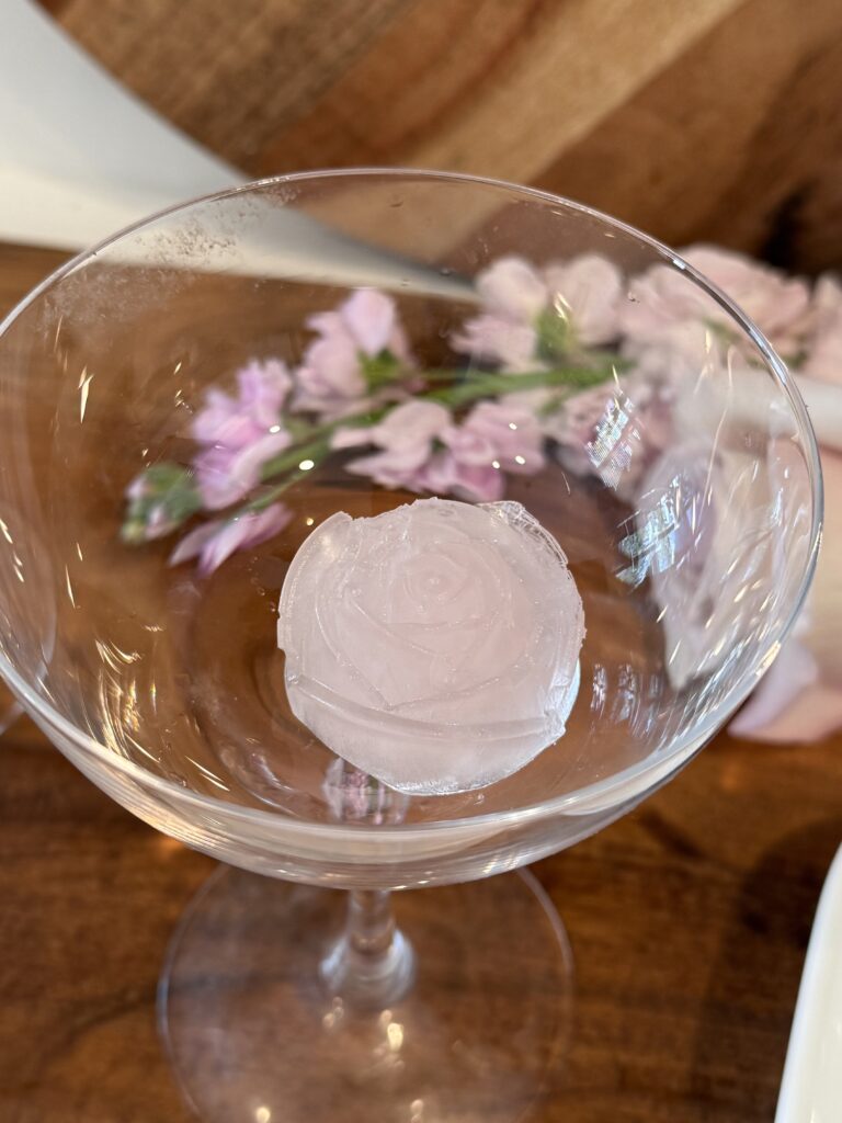 Rose ice cube shape in a coupe glass on a cutting board on the counter, with flowers in the background.