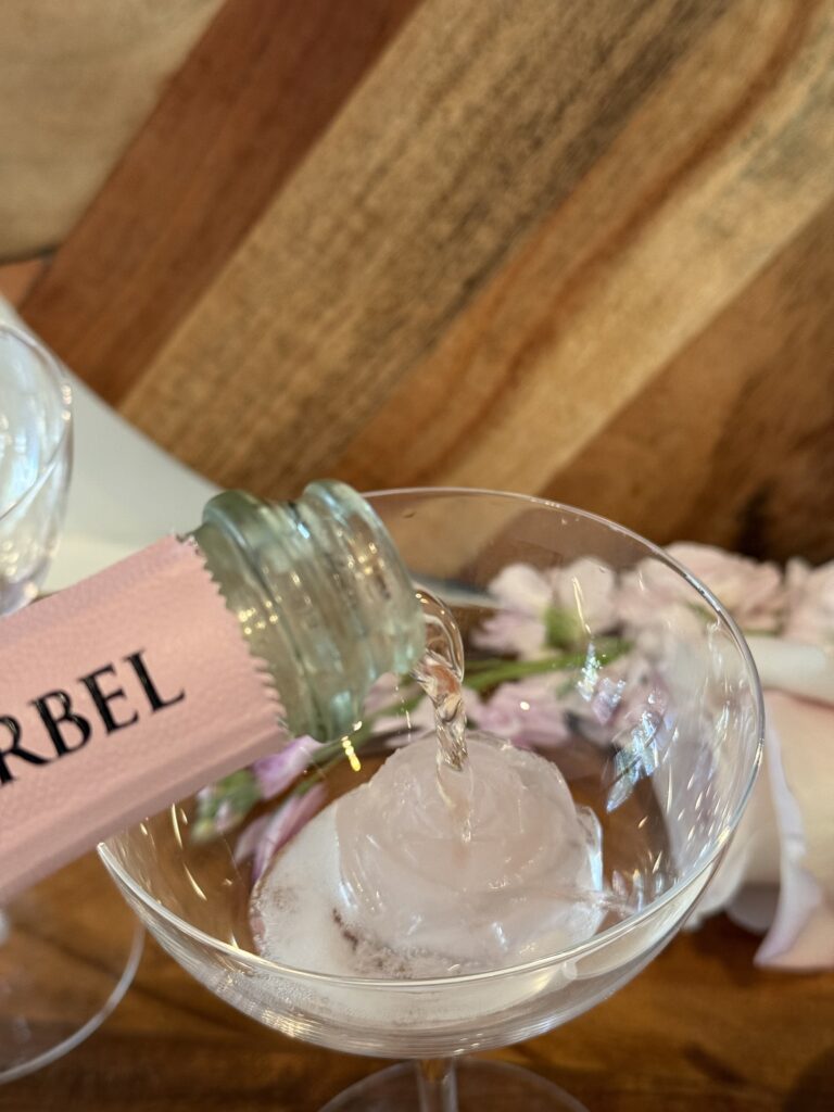 Pouring brut rose champagne into a coupe glass on a brown cutting board.