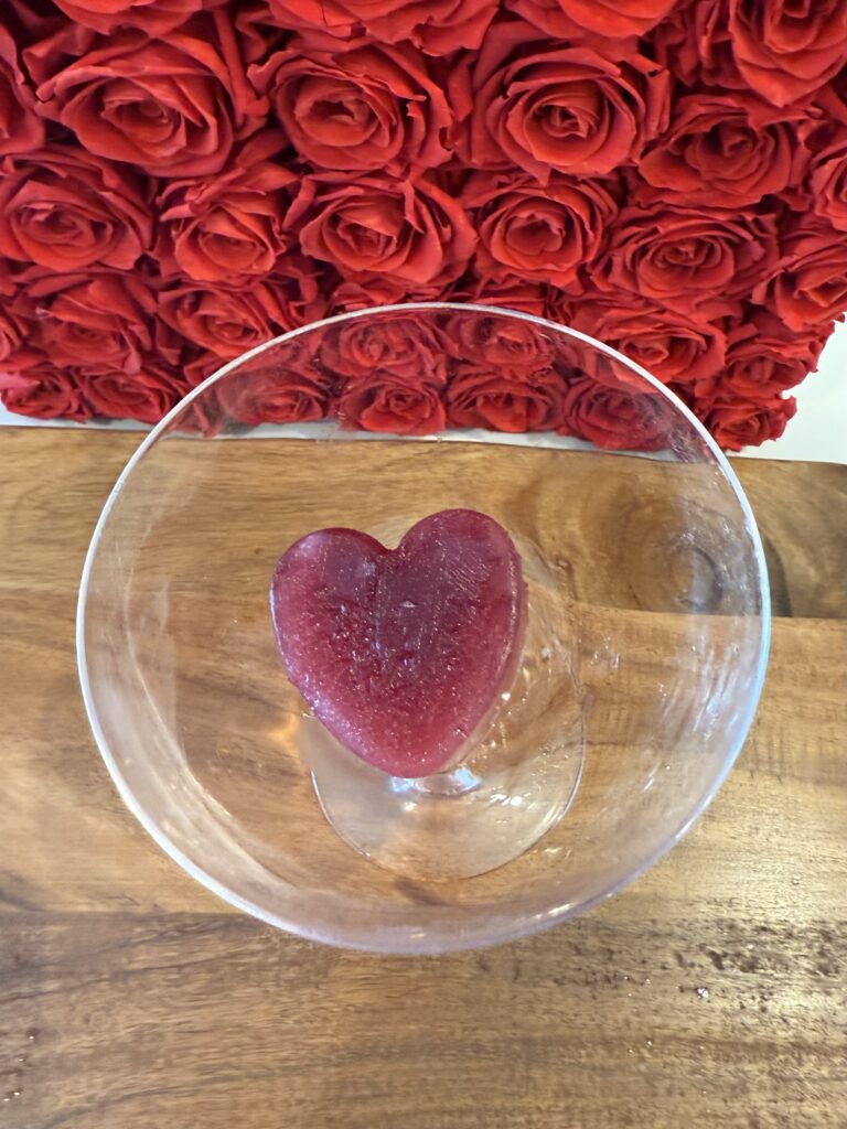 Heart shape ice cube in a coupe glass on a cutting board on the counter.