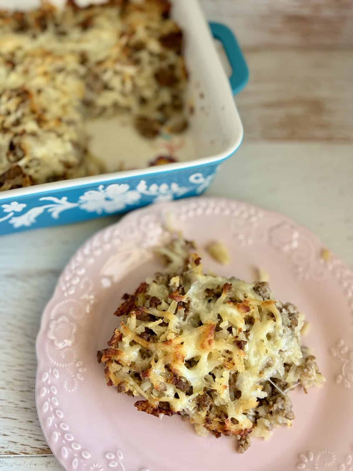 Close-up of a slice of sausage potato casserole on a pink plate, showing melted cheese, sausage, and crispy topping.