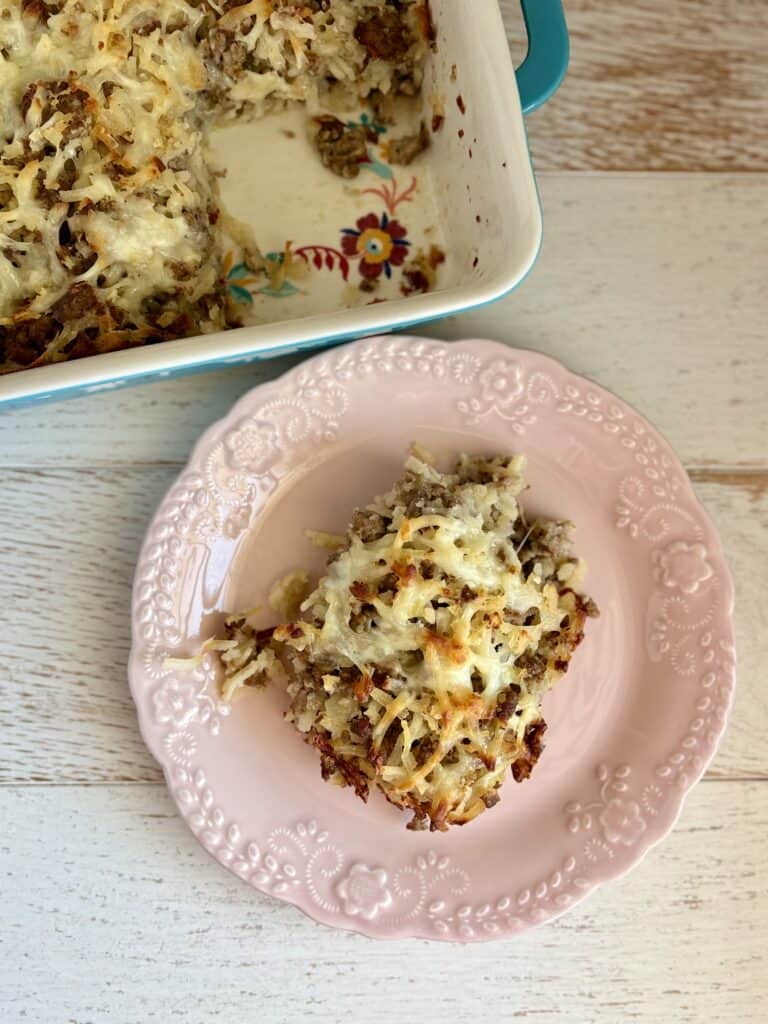 A slice of sausage potato casserole on a pink plate with melted cheese and cooked sausage pieces, served from a baking dish.
