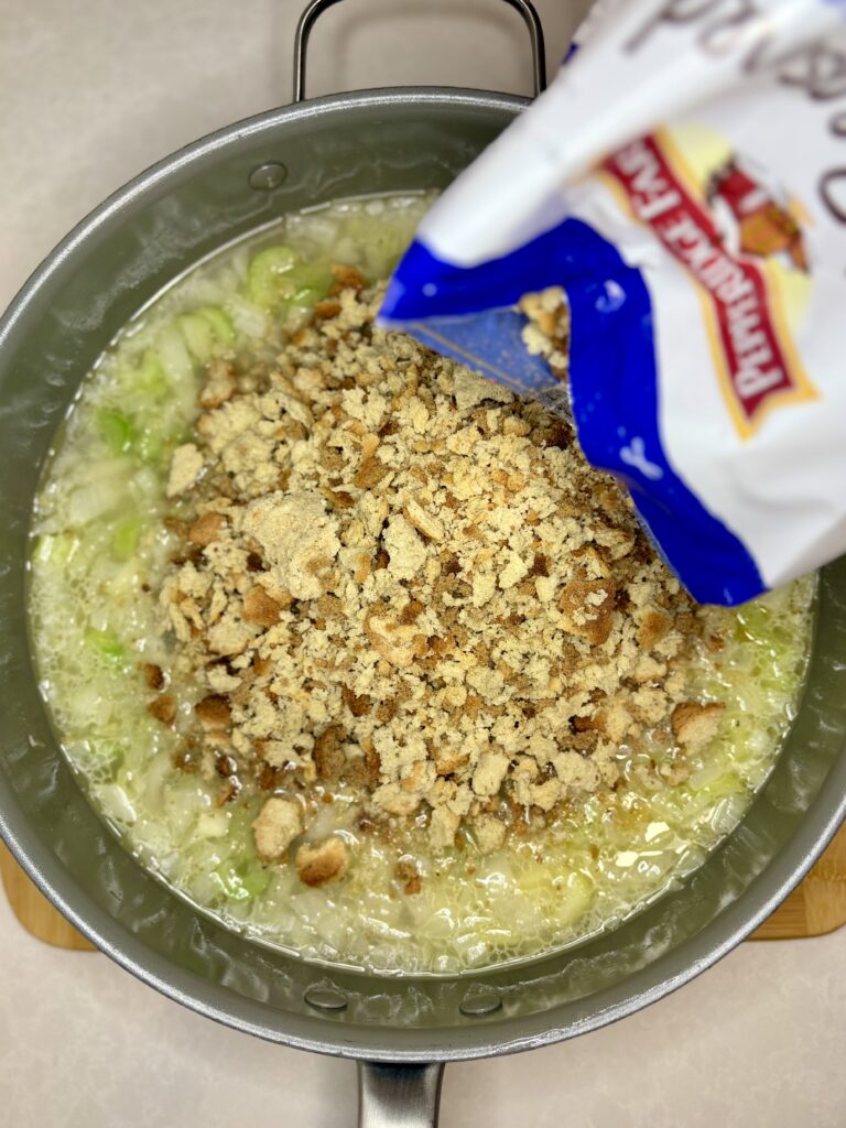 a pan on a counter with broth, celery and  onion with stuffing mix being poured iin