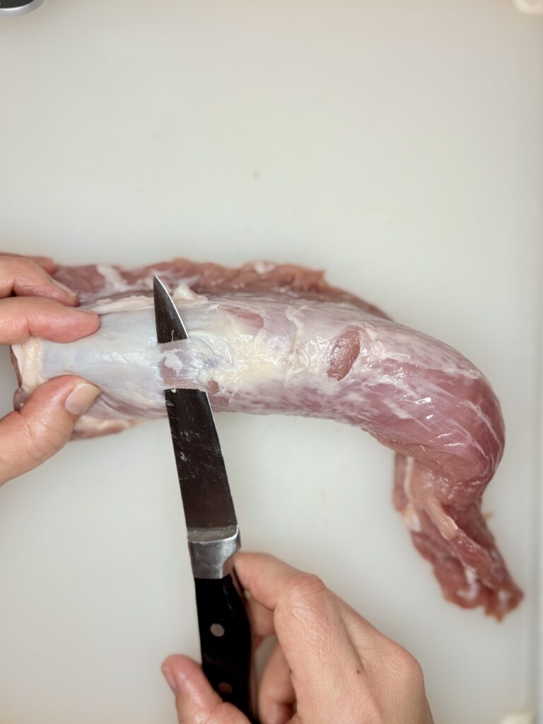 a knife sliding under and cutting the silver tendon from a pork tenderloin
