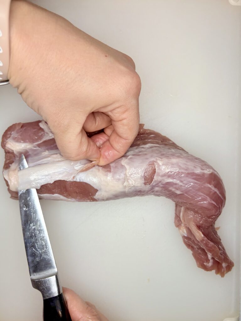 a piece of a silver tendon being removed from pork tenderloin with a knife