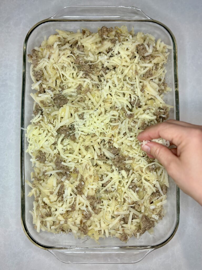 sausage potato mixture in a casserole dish being topped with shredded mozzarella cheese 