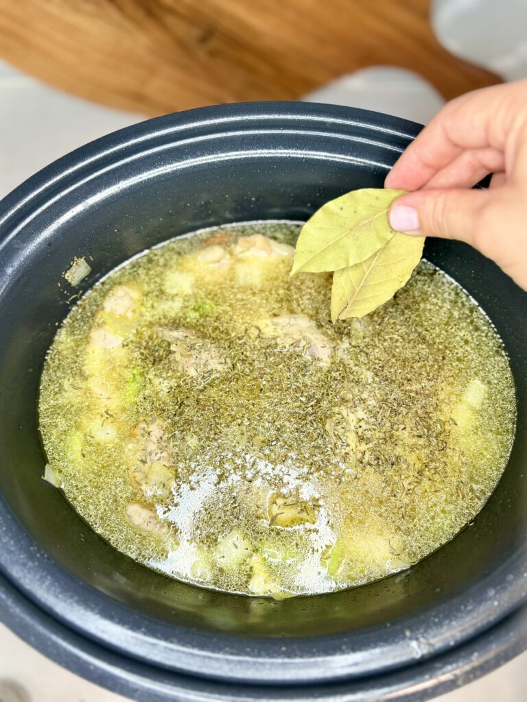 bay leaves and seasoning being added to slow cooker with broth, chicken and vegetables