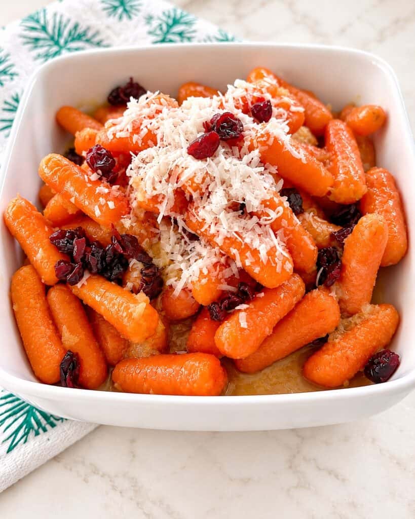 Pineapple glazed baby carrots topped with shredded coconut and dried cranberries in a white serving bowl.”