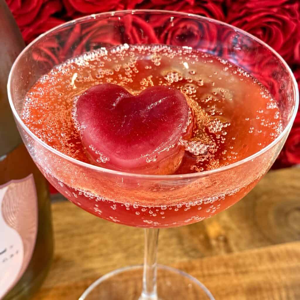 Valentine's Day cocktail with a heart-shaped ice cube in a glass, featuring a pinkish-red hue and bubbles, on a romantic red rose background.