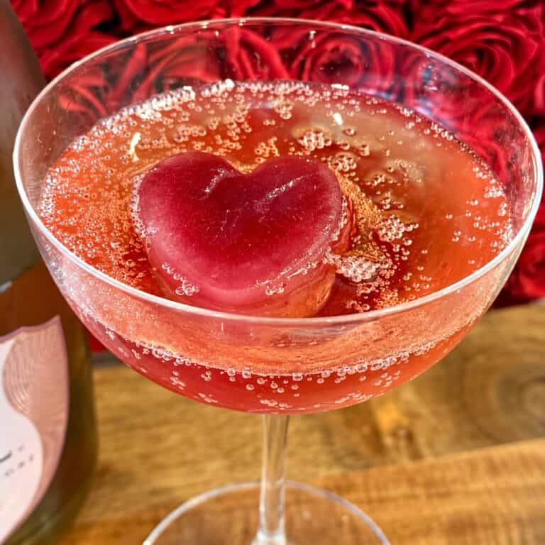 Valentine's Day cocktail with a heart-shaped ice cube in a glass, featuring a pinkish-red hue and bubbles, on a romantic red rose background.