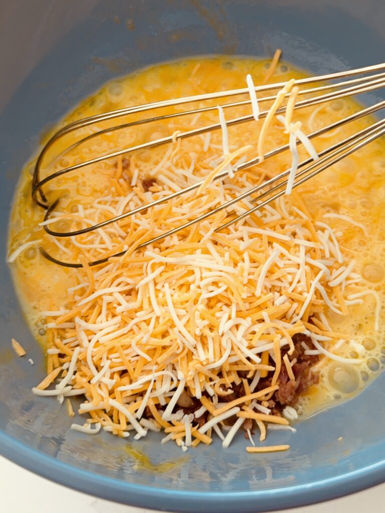 whisked eggs with shredded Colby jack cheese and bacon pieces in a blue mixing bowl on the counter.