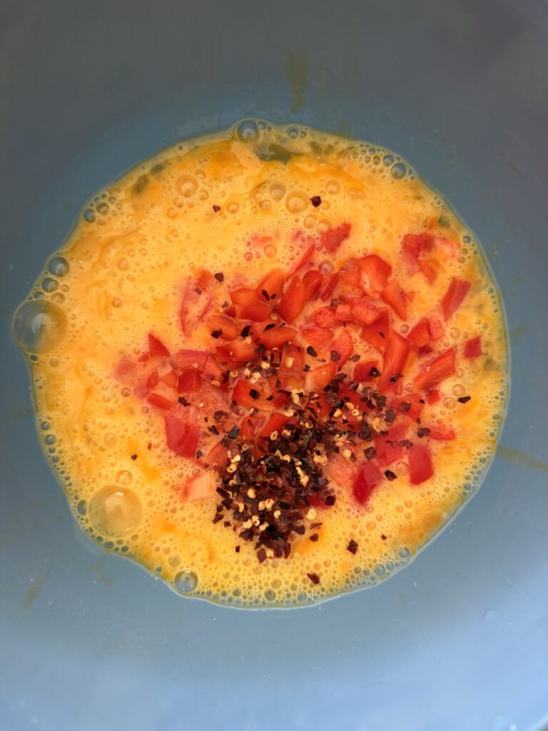 whisked eggs, chopped red pepper and red pepper flakes in a blue mixing bowl on the counter.