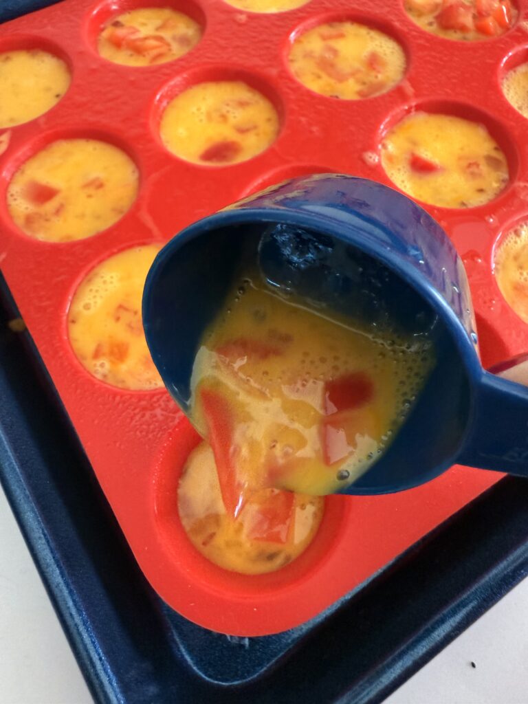 pouring g mixing egg batter into mini muffin pan on a dark blue baking sheet on the counter .