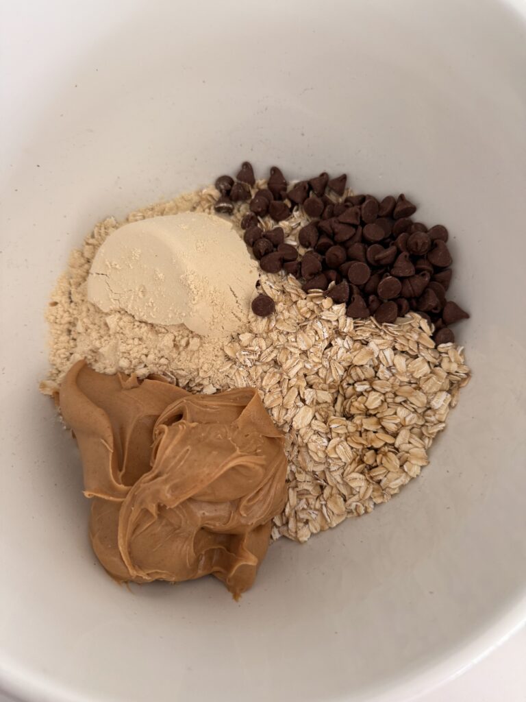 rolled oats, peanut butter, maple syrup, vanilla protein powder, semi-sweet chocolate chips in a large white mixing bowl on the counter.