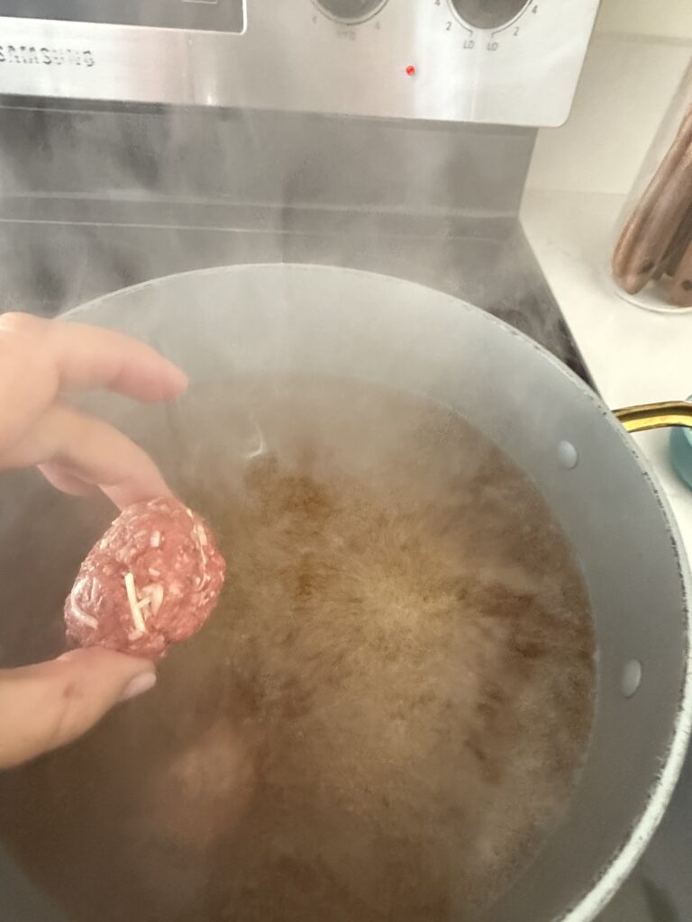 A meatball being placed from a blue measuring cup into the broth in a large pot on the stovetop.