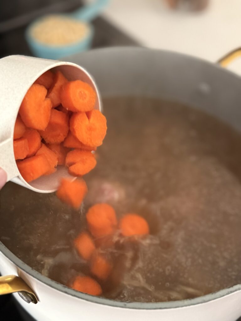 chopped carrots being poured from a blue measuring cup into the broth in a large pot on the stovetop.