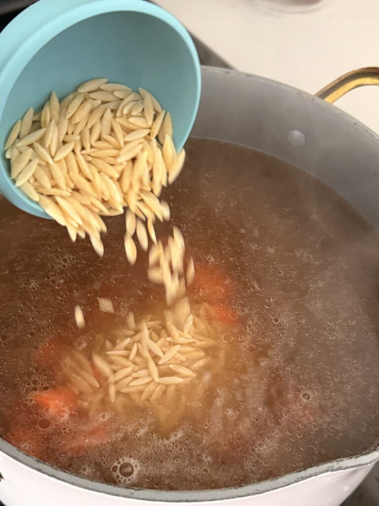 orzo pasta being poured from a blue measuring cup into the broth in a large pot on the stovetop.