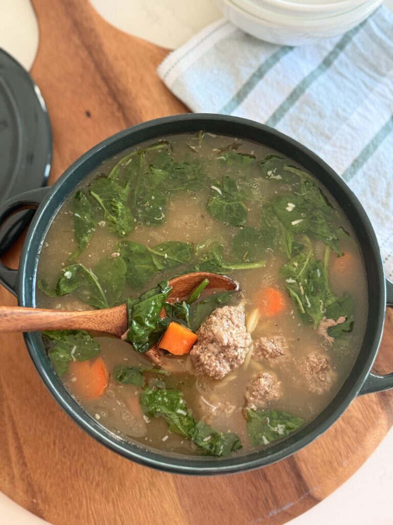 Italian wedding soup in a large green pot on a brown cutting board on the counter.