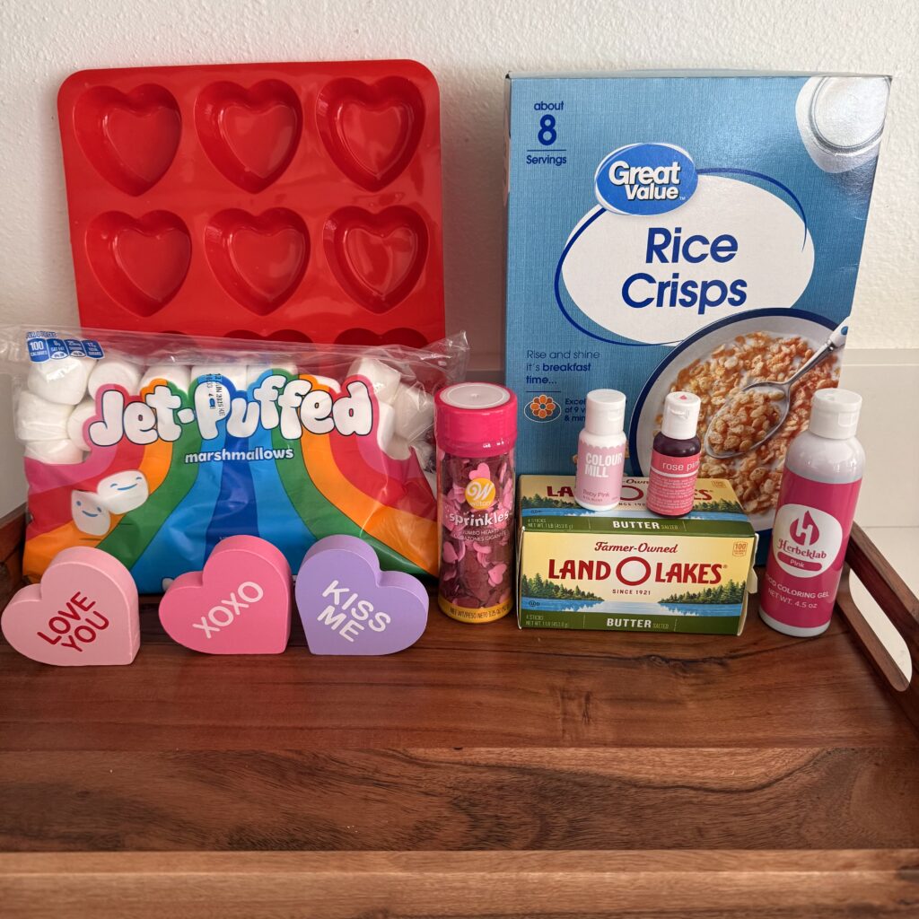 Rice crips cereal, large marshmallows, edible hearts, heart-shaped mold, butter, pink food colorings, and valentine decorations. On a brown tray on the counter.