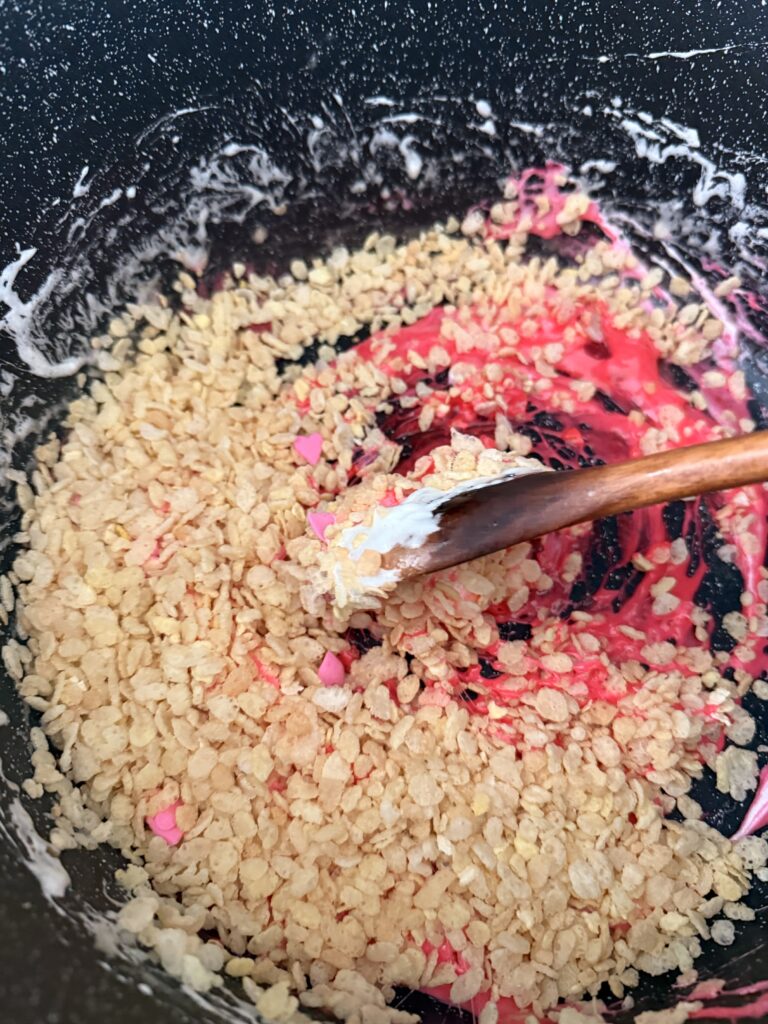 Edible sugar hearts with rice crispy cereal and pink marshmallow mixture are stirred by a wooden spoon in a large black pot on the stovetop.