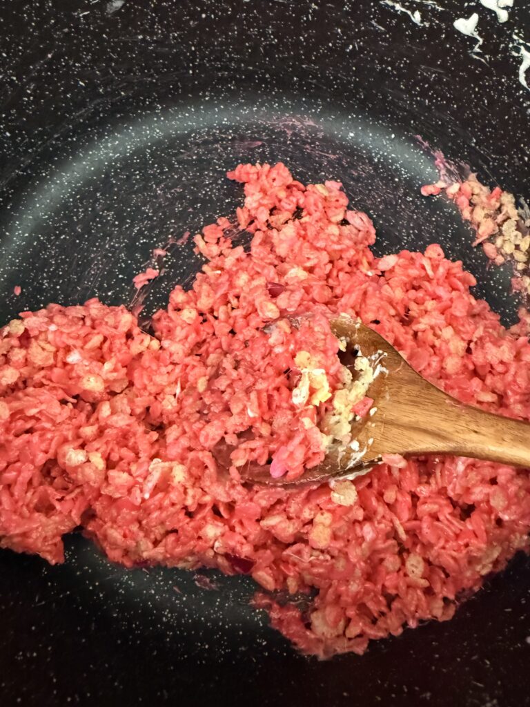 Edible sugar hearts with rice crispy cereal and pink marshmallow mixture are stirred by a wooden spoon in a large black pot on the stovetop.