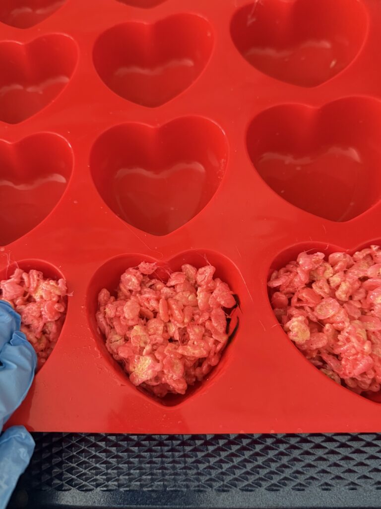 Using food-greased food gloves press pink rice crispy treats into heart-shaped molds.