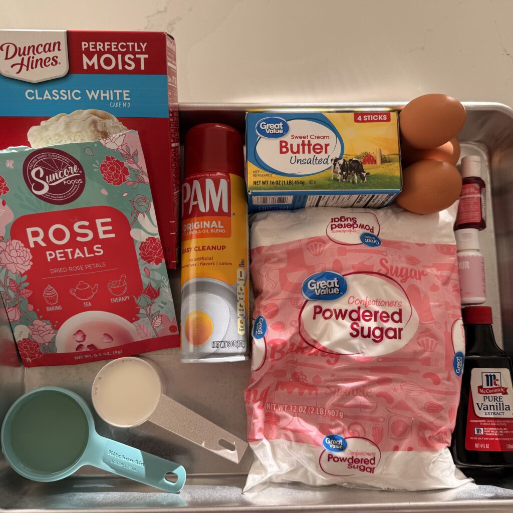 White cake mix box, unsalted butter, eggs, non-stick spray, milk in a measuring cup, vegetable oil in a measuring cup, powdered sugar, vanilla extract, and edible rose petals on a silver baking tray. On the counter.