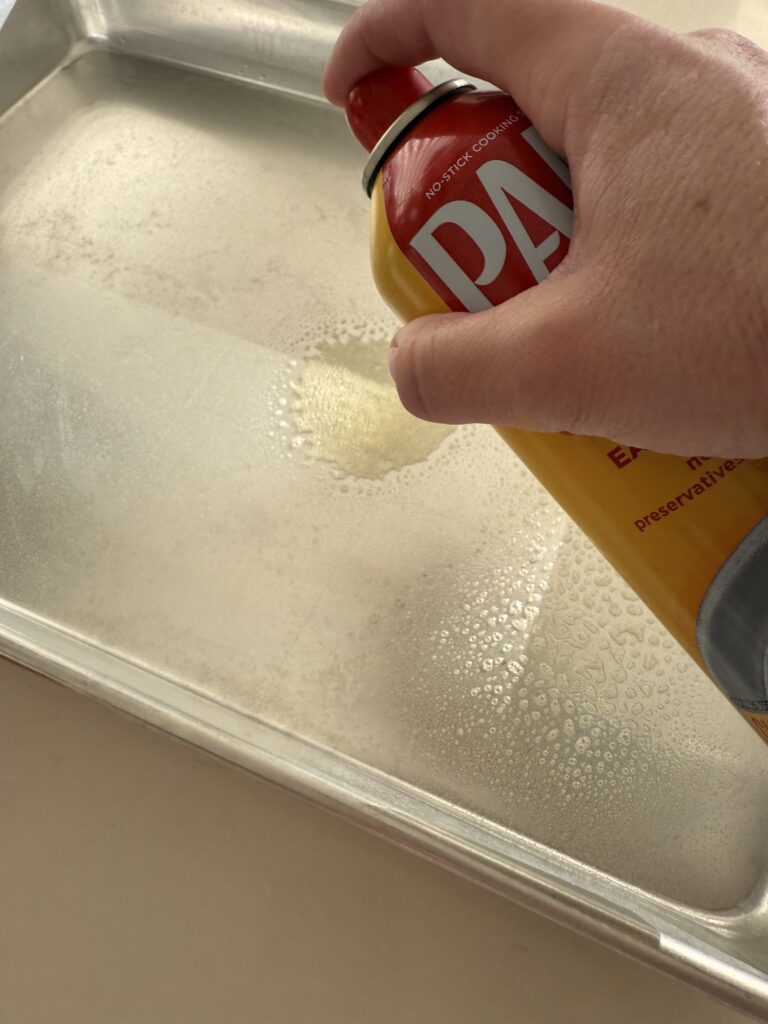 Spraying non-stick cooking spray onto an 18x13 inch rimmed cake pan on the counter.