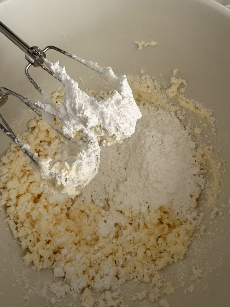 An electric hand mixing blades and a large white bowl with butter and powdered sugar on the counter.