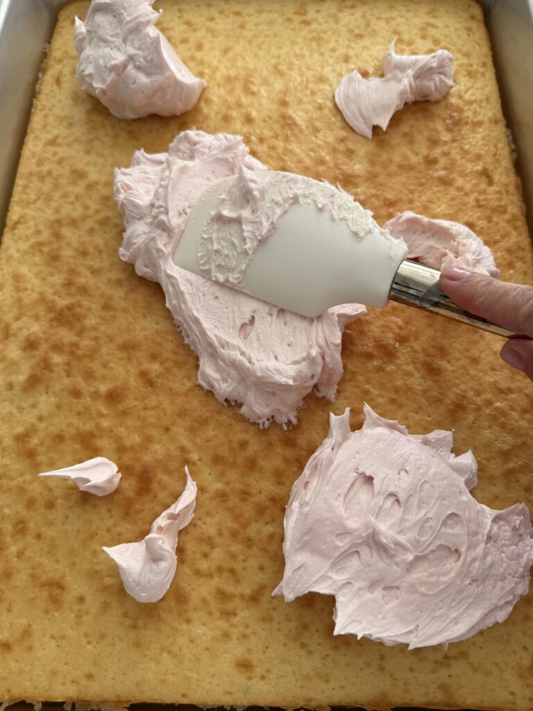 The back of a spatula spreads the pink icing onto the cake in a silver 18x13-inch cake pan on the counter.